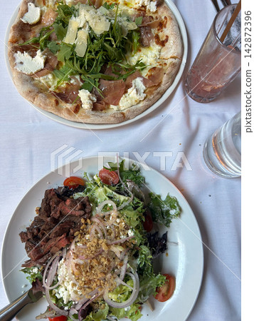 Prosciutto arugula pizza and steak salad on a white tablecloth Prosciutto arugula pizza and steak salad on a white tablecloth 142872396