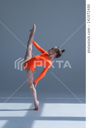 A little Caucasian girl gymnast in an orange leotard demonstrates her skills on a white background. A little Caucasian girl gymnast in an orange leotard demonstrates her skills on a white background. 142872486