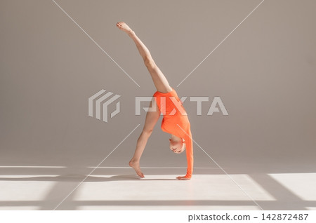 A little Caucasian girl gymnast in an orange leotard demonstrates her skills on a white background. A little Caucasian girl gymnast in an orange leotard demonstrates her skills on a white background. 142872487