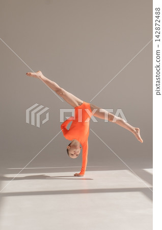 A little Caucasian girl gymnast in an orange leotard demonstrates her skills on a white background. A little Caucasian girl gymnast in an orange leotard demonstrates her skills on a white background. 142872488