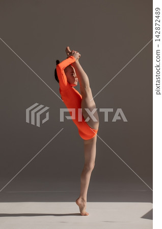 A little Caucasian girl gymnast in an orange leotard demonstrates her skills on a white background. A little Caucasian girl gymnast in an orange leotard demonstrates her skills on a white background. 142872489