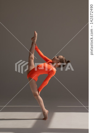 A little Caucasian girl gymnast in an orange leotard demonstrates her skills on a white background. A little Caucasian girl gymnast in an orange leotard demonstrates her skills on a white background. 142872490