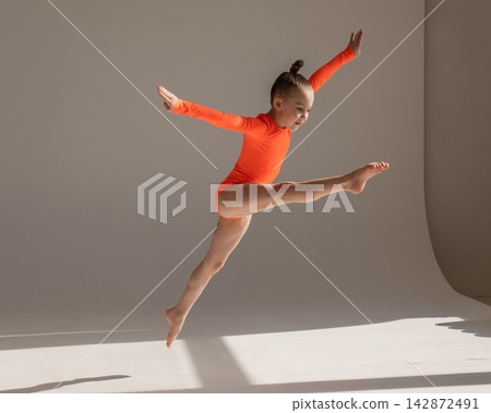 A little Caucasian girl gymnast in an orange leotard demonstrates her skills on a white background. A little Caucasian girl gymnast in an orange leotard demonstrates her skills on a white background. 142872491