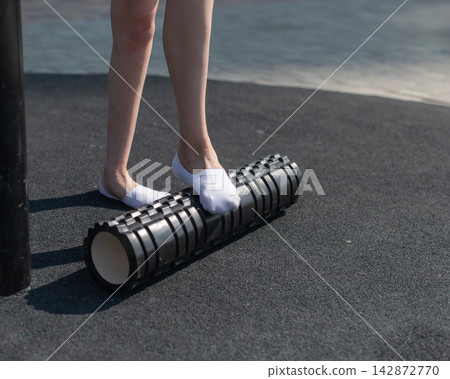 A Caucasian woman uses a foam roller to stretch her feet on a sports field. Close-up of her feet. A Caucasian woman uses a foam roller to stretch her feet on a sports field. Close-up of her feet. 142872770
