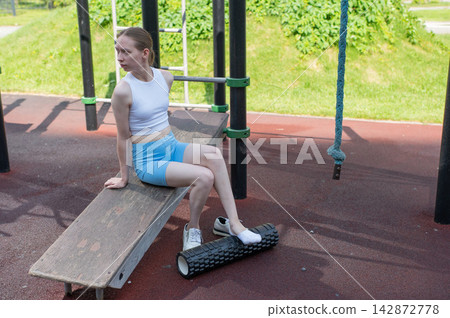 A Caucasian woman stretches her feet with a foam roller at a sports ground. A Caucasian woman stretches her feet with a foam roller at a sports ground. 142872778