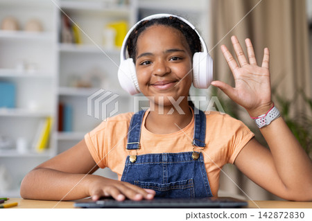 Smiling Black teenage girl wearing headphones waves toward the camera at home 142872830