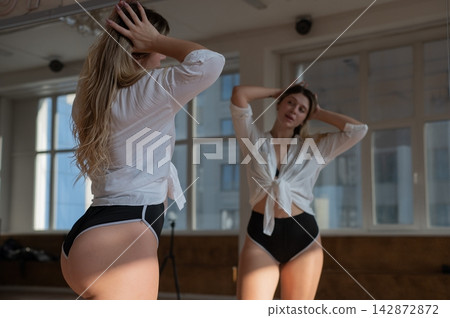 A Caucasian woman in short shorts poses in front of a mirror in a dance class. A Caucasian woman in short shorts poses in front of a mirror in a dance class. 142872872