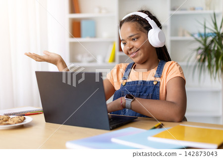 Black teenage girl wearing headphones gestures while using a laptop at home 142873043