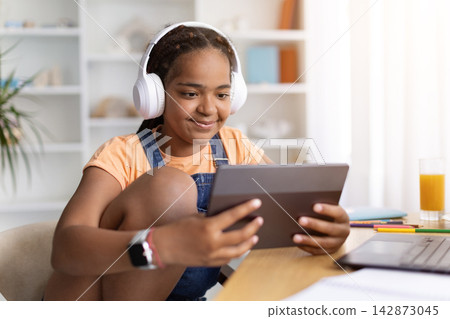 Black teenage girl wearing headphones using tablet at desk at home, Black teenage girl wearing headphones using tablet at desk at home, 142873045
