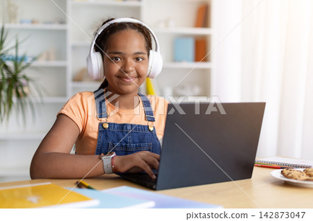 Smiling Black teenage girl wearing headphones sits behind a laptop at home Smiling Black teenage girl wearing headphones sits behind a laptop at home 142873047