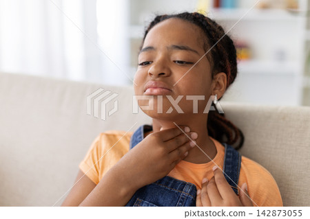 Black teenage girl touching her throat while sitting at home, showing discomfort Black teenage girl touching her throat while sitting at home, showing discomfort 142873055