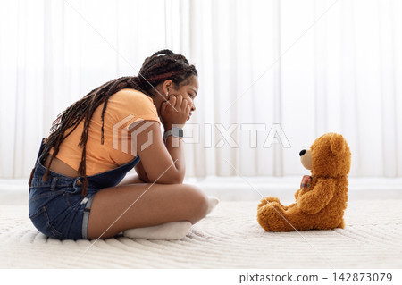 Thoughtful Black girl sitting on floor, looking at teddy bear at home Thoughtful Black girl sitting on floor, looking at teddy bear at home 142873079