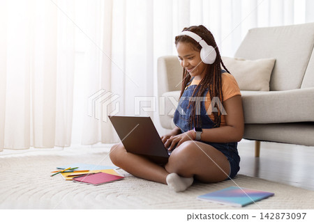 Black teenage girl wearing headphones smiling while using laptop at home Black teenage girl wearing headphones smiling while using laptop at home 142873097