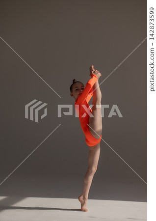 A little Caucasian girl gymnast in an orange leotard demonstrates her skills on a white background. A little Caucasian girl gymnast in an orange leotard demonstrates her skills on a white background. 142873599