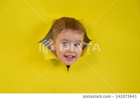 A mischievous boy peeks out through a hole in a yellow cardboard background. A mischievous boy peeks out through a hole in a yellow cardboard background. 142873643