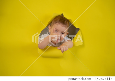 A mischievous boy peeks through a hole in a yellow paper background and gives a thumbs up. A mischievous boy peeks through a hole in a yellow paper background and gives a thumbs up. 142874112