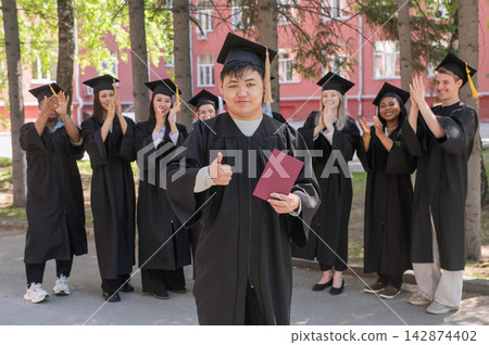 Young Asian man holds diploma while classmates applaud her. Young Asian man holds diploma while classmates applaud her. 142874402