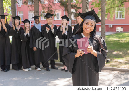 Young African woman holds diploma while classmates applaud her.  142874403