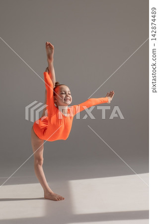 A little Caucasian girl gymnast in an orange leotard demonstrates her skills on a white background. A little Caucasian girl gymnast in an orange leotard demonstrates her skills on a white background. 142874489