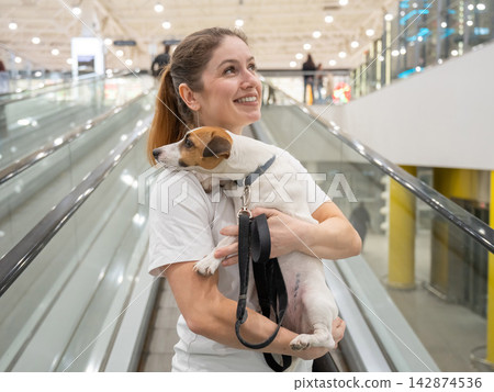A Caucasian woman holds a dog on a shopping mall conveyor belt. Dog friendly environment. A Caucasian woman holds a dog on a shopping mall conveyor belt. Dog friendly environment. 142874536