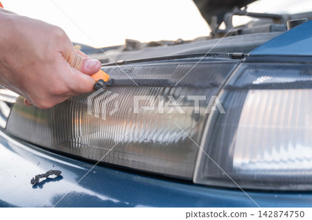 A man removes double-sided tape from a car headlight. A man removes double-sided tape from a car headlight. 142874750