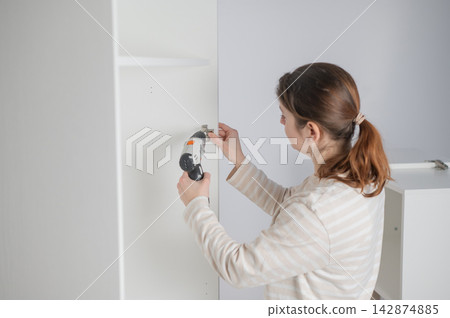 A Caucasian woman uses a screwdriver to tighten a screw while assembling a cabinet. 142874885