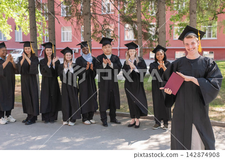Young Caucasian man holding diploma while classmates applaud him. Young Caucasian man holding diploma while classmates applaud him. 142874908