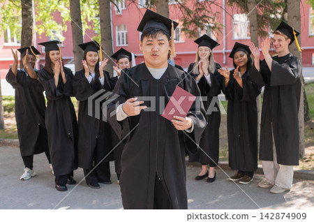 Young Asian man holds diploma while classmates applaud her. Young Asian man holds diploma while classmates applaud her. 142874909