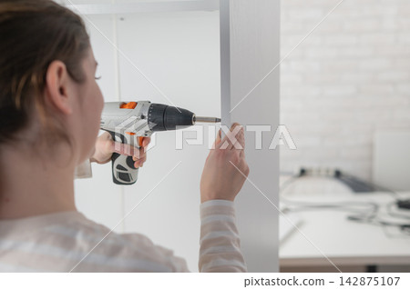 A Caucasian woman uses a screwdriver to tighten a screw while assembling a cabinet. A Caucasian woman uses a screwdriver to tighten a screw while assembling a cabinet. 142875107