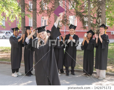 Young Caucasian woman holding diploma while classmates applaud her. Young Caucasian woman holding diploma while classmates applaud her. 142875172