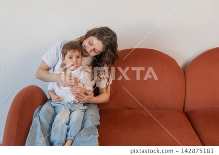 A Caucasian woman and her two-year-old son hug while sitting on a sofa. A Caucasian woman and her two-year-old son hug while sitting on a sofa. 142875181