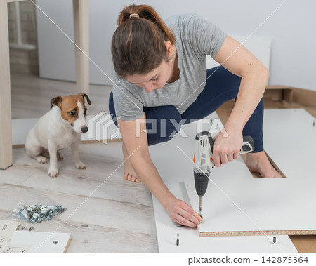 A Caucasian woman uses a screwdriver to tighten a screw while assembling a cabinet. A Caucasian woman uses a screwdriver to tighten a screw while assembling a cabinet. 142875364