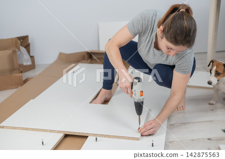 A Caucasian woman uses a screwdriver to tighten a screw while assembling a cabinet. A Caucasian woman uses a screwdriver to tighten a screw while assembling a cabinet. 142875563