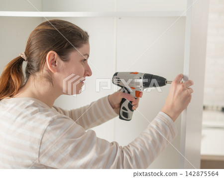 A Caucasian woman uses a screwdriver to tighten a screw while assembling a cabinet. A Caucasian woman uses a screwdriver to tighten a screw while assembling a cabinet. 142875564