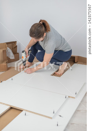 A Caucasian woman uses a screwdriver to tighten a screw while assembling a cabinet. A Caucasian woman uses a screwdriver to tighten a screw while assembling a cabinet. 142875567