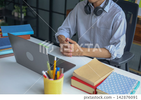 A man listening to a lesson during a virtual classroom remote learning session with laptop and books on the table. 142876091