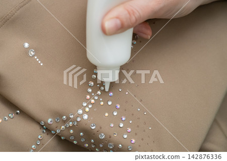 A woman decorates a gymnastics costume with rhinestones. Rhythmic gymnastics. Glue in a tube. A woman decorates a gymnastics costume with rhinestones. Rhythmic gymnastics. Glue in a tube. 142876336