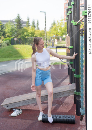 A Caucasian woman stretches her feet with a foam roller at a sports ground. A Caucasian woman stretches her feet with a foam roller at a sports ground. 142876484
