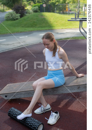 A Caucasian woman stretches her feet with a foam roller at a sports ground. 142876486