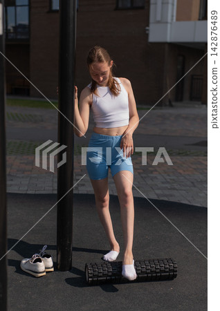 A Caucasian woman stretches her feet with a foam roller at a sports ground. A Caucasian woman stretches her feet with a foam roller at a sports ground. 142876489