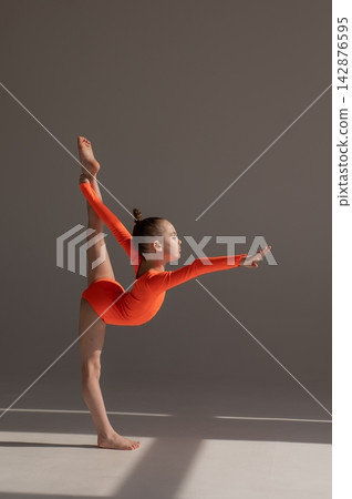 A little Caucasian girl gymnast in an orange leotard demonstrates her skills on a white background. A little Caucasian girl gymnast in an orange leotard demonstrates her skills on a white background. 142876595