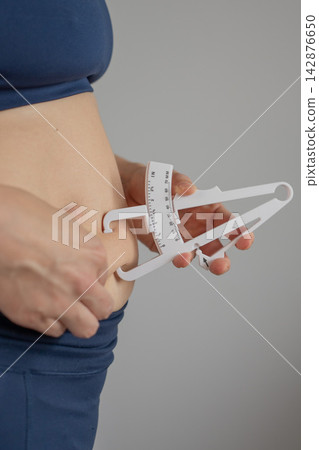 A woman measures the amount of subcutaneous fat on her abdomen using a caliper. A woman measures the amount of subcutaneous fat on her abdomen using a caliper. 142876650