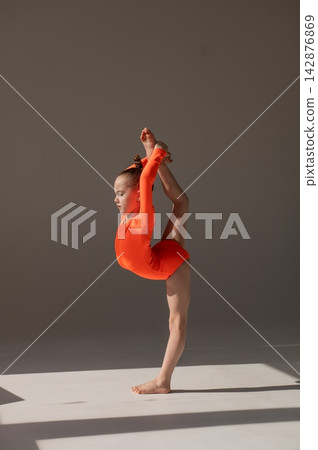 A little Caucasian girl gymnast in an orange leotard demonstrates her skills on a white background. A little Caucasian girl gymnast in an orange leotard demonstrates her skills on a white background. 142876869
