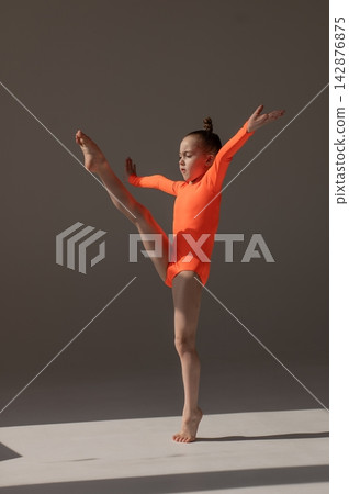 A little Caucasian girl gymnast in an orange leotard demonstrates her skills on a white background. A little Caucasian girl gymnast in an orange leotard demonstrates her skills on a white background. 142876875