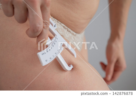 A woman measures the amount of subcutaneous fat on her thigh using calipers. A woman measures the amount of subcutaneous fat on her thigh using calipers. 142876878