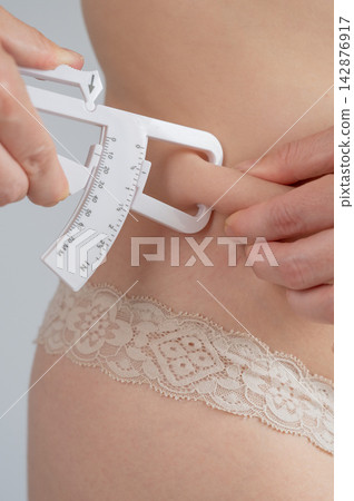 A woman measures the amount of subcutaneous fat on her abdomen using a caliper. A woman measures the amount of subcutaneous fat on her abdomen using a caliper. 142876917