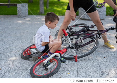 A Caucasian woman is riding a bicycle with her son. The boy fell, and the mother feels sorry for him. A Caucasian woman is riding a bicycle with her son. The boy fell, and the mother feels sorry for him. 142876927