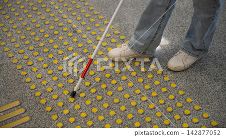 A blind woman walks on tactile paving. Close-up of her feet and a walking stick. A blind woman walks on tactile paving. Close-up of her feet and a walking stick. 142877052