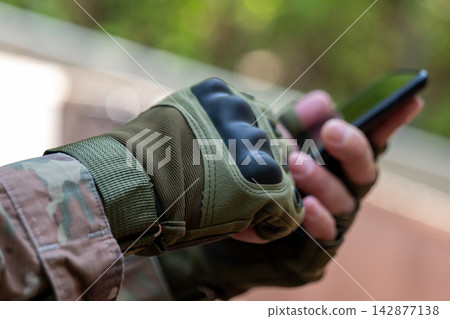 Gloved military hand holding smartphone outdoors in soft daylight Gloved military hand holding smartphone outdoors in soft daylight 142877138