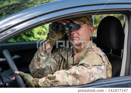 Camouflaged man observing through binoculars while sitting inside a car Camouflaged man observing through binoculars while sitting inside a car 142877147